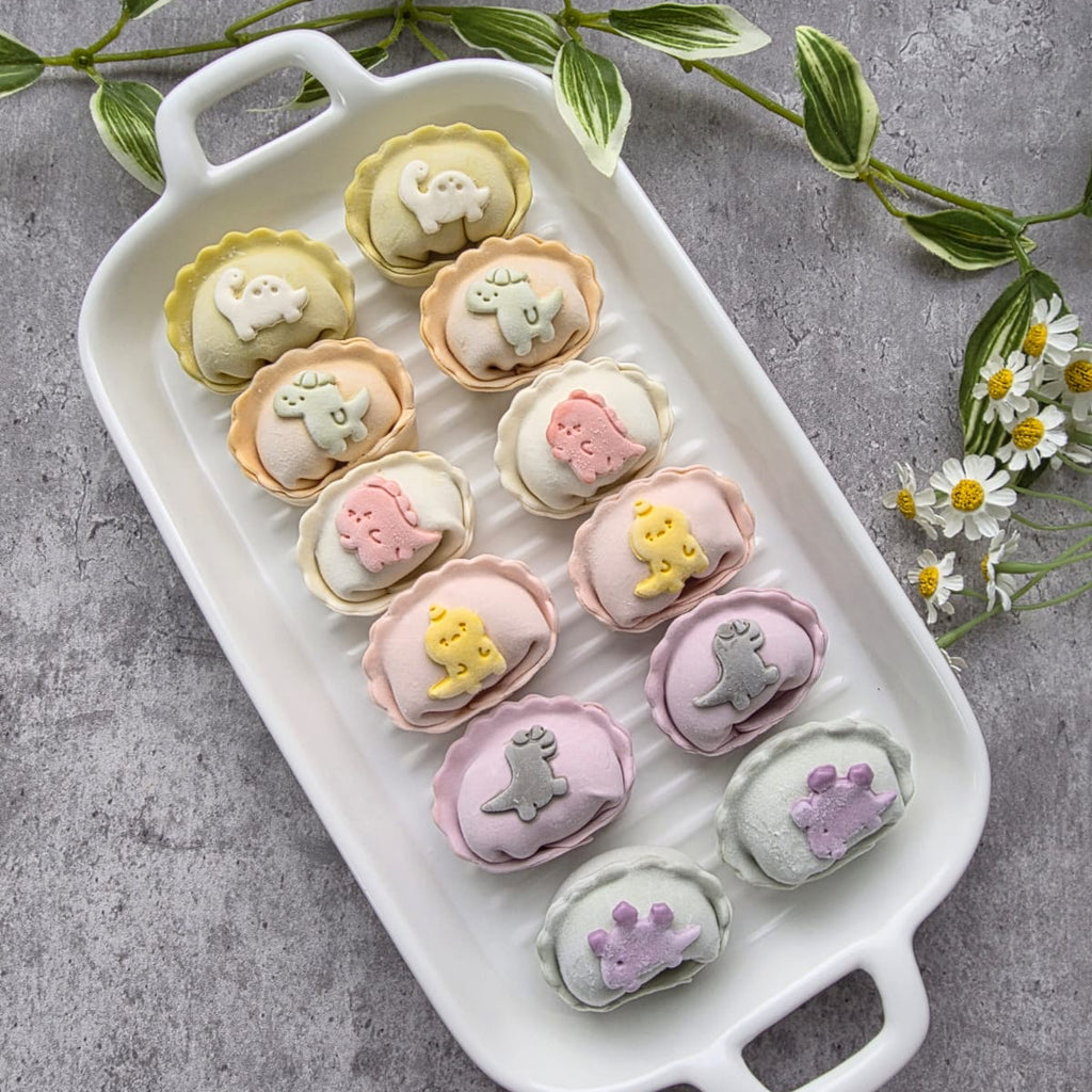 Decorative dumplings with dino shapes on a white tray on a gray surface with green leaves and flowers.
