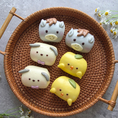 Six cute, cartoon-shaped steamed buns arranged in a round woven wicker tray on a gray stone surface.